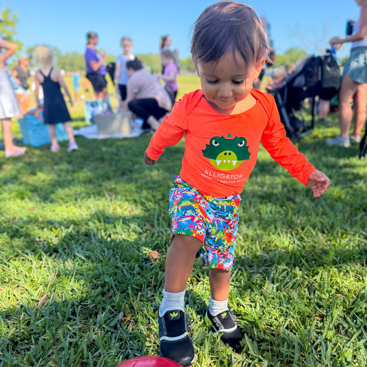 Child in a ShapeShifter Fish and Friends orange UPF onesie and colorful shorts playing on grass with other people in the background