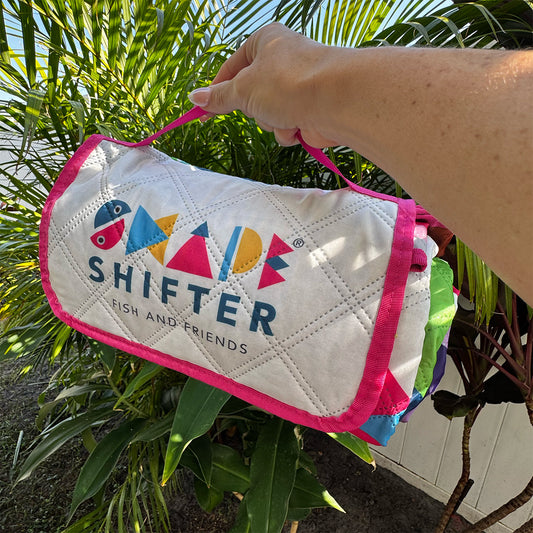 Hand holding a Shifter Fish and Friends rolled up beach matt with colorful design against a green leafy background