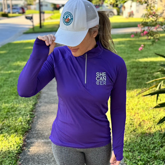 Woman wearing a purple long-sleeve shirt with 'SHE CAN CER' text, white cap, and gray leggings outdoors, for cancer patients, chemotherapy, radiation