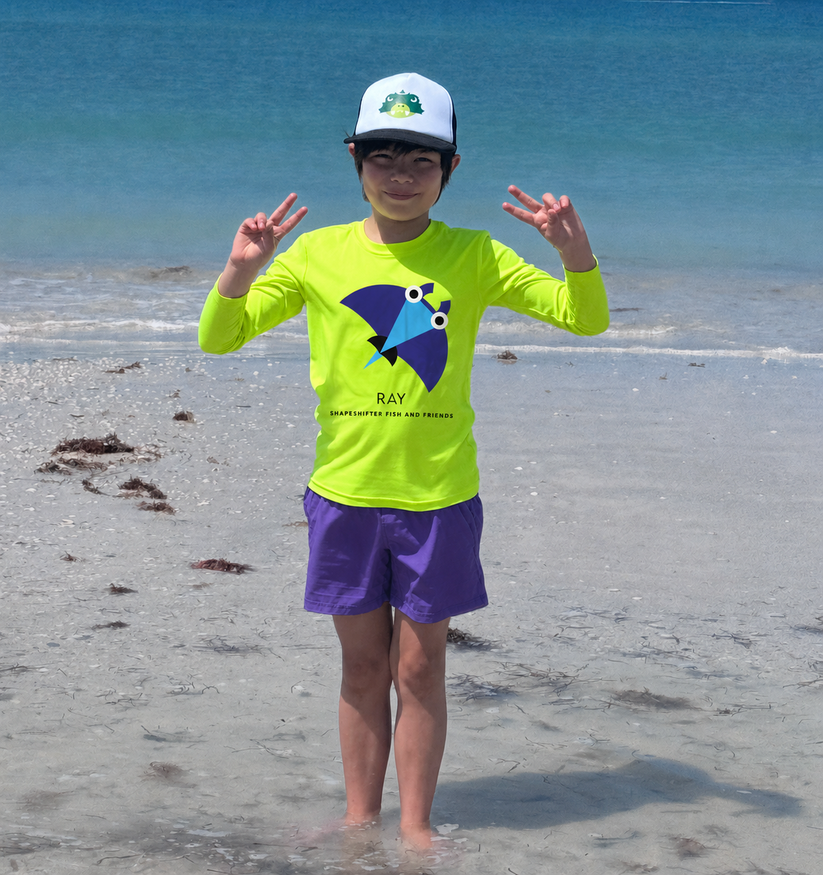 A child standing on a sandy beach in shallow water, wearing a bright neon yellow long-sleeve swim shirt with a blue and yellow graphic, purple swim shorts, and a white cap.