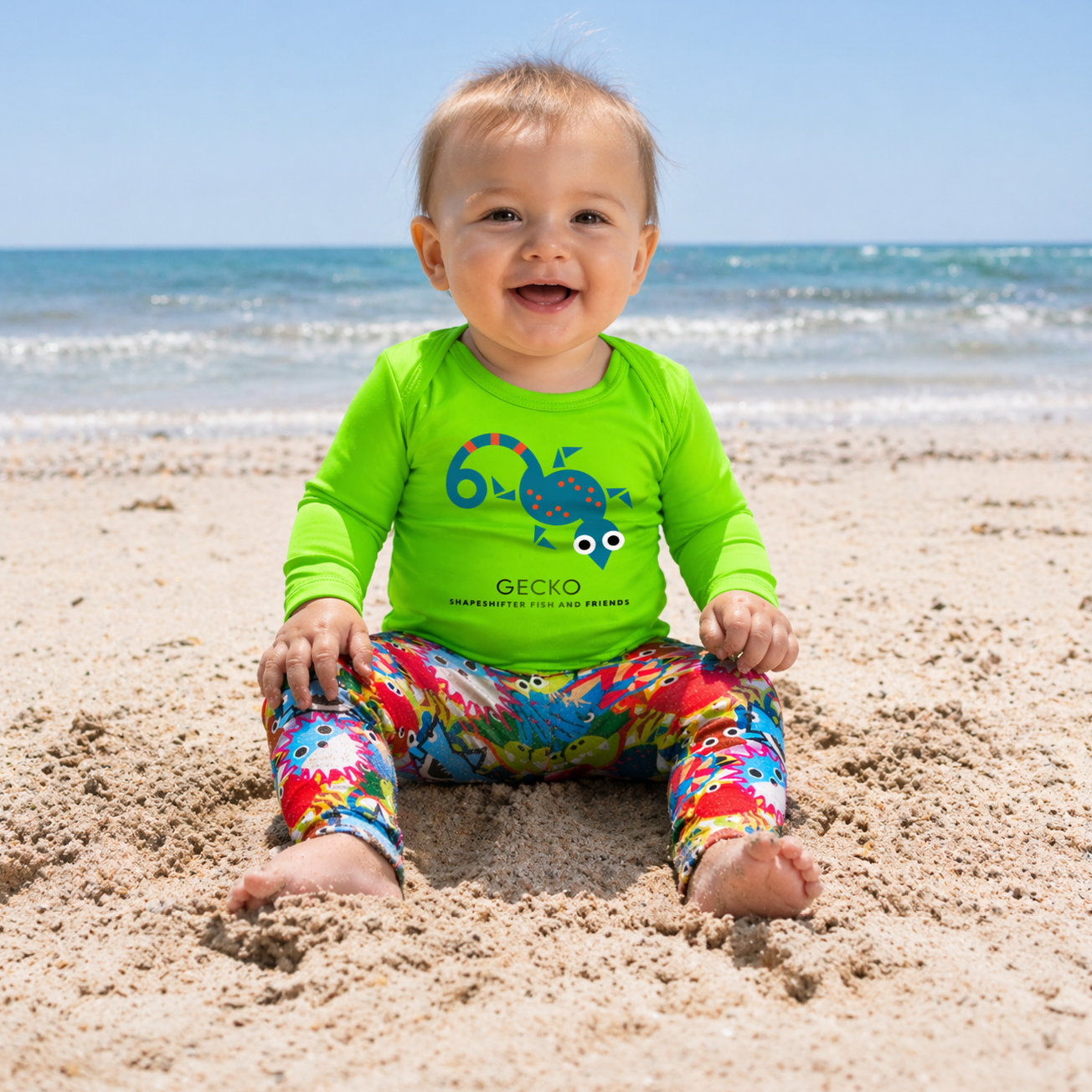 Baby wearing a green upf onesie with a gecko design and colorful sun protective swim pants sitting on a sandy beach.