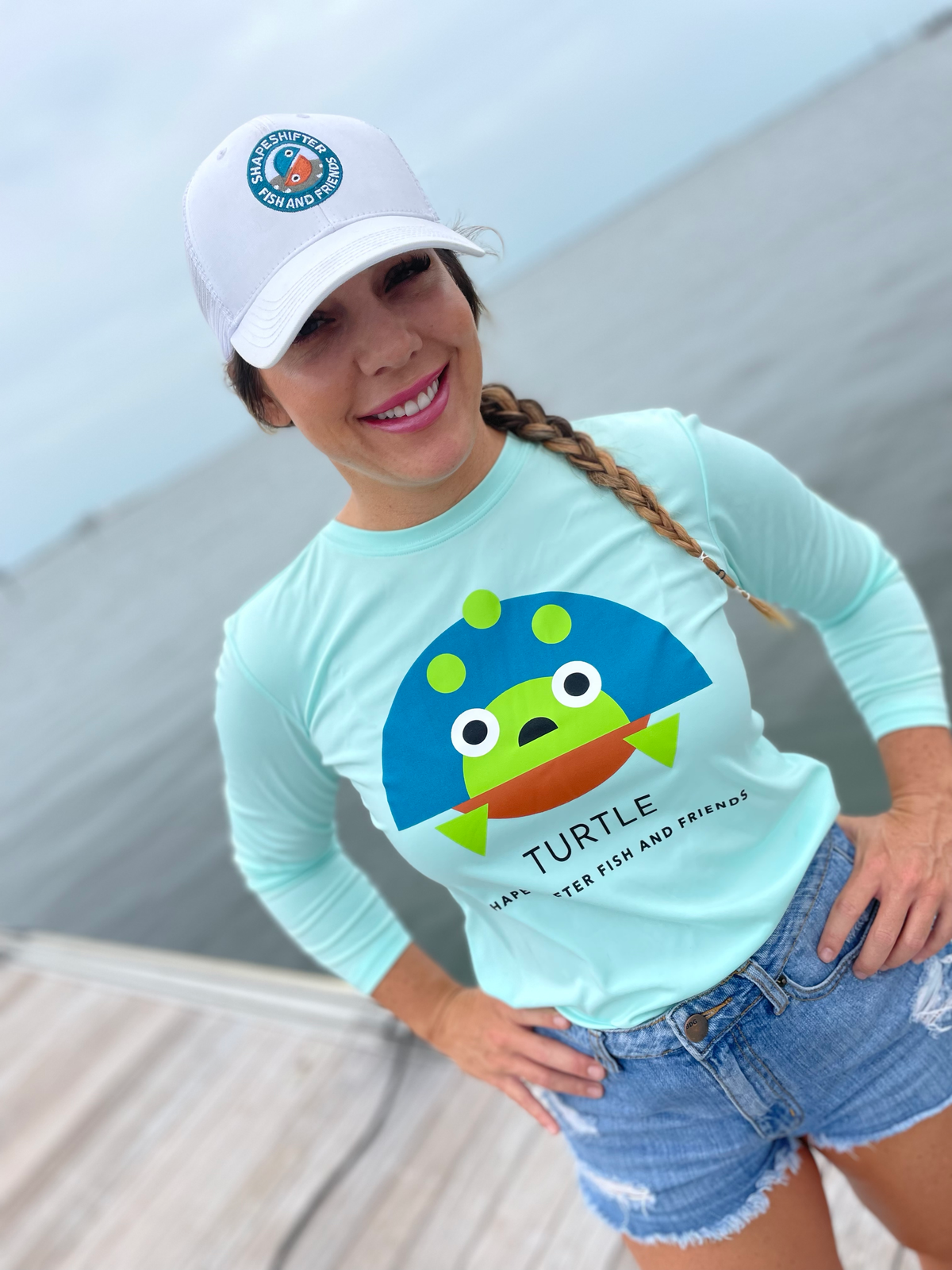 Woman wearing a Upf50 Sun Protection Shirt by ShapeShifter Fish and Friends with a turtle graphic and a white cap on a boat dock.