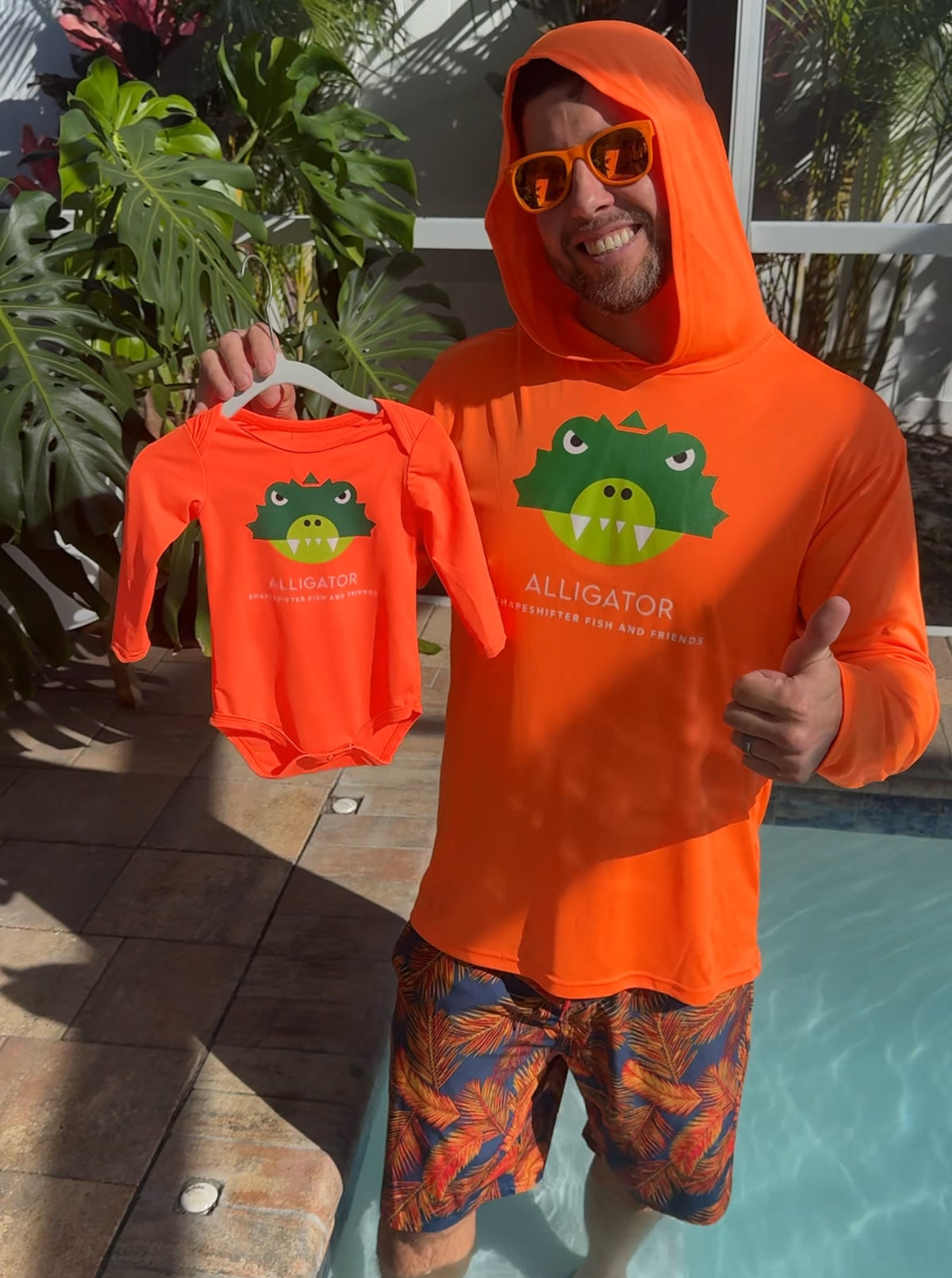 Man wearing a Neon Orange Alligator Hooded Sun Protective Shirt UPF 50+ by ShapeShifter Fish and Friends, holding a matching alligator baby onesie by a pool.