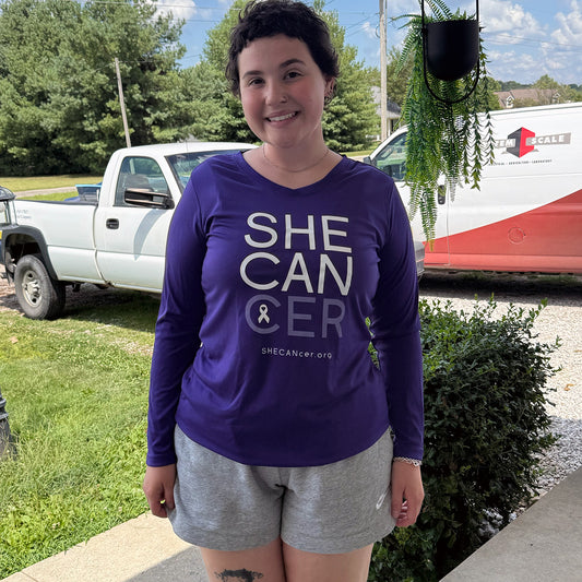 Woman wearing a purple 'SHECANCER' UPF shirt by ShapeShifter Fish and Friends standing outdoors with vehicles and greenery in the background.