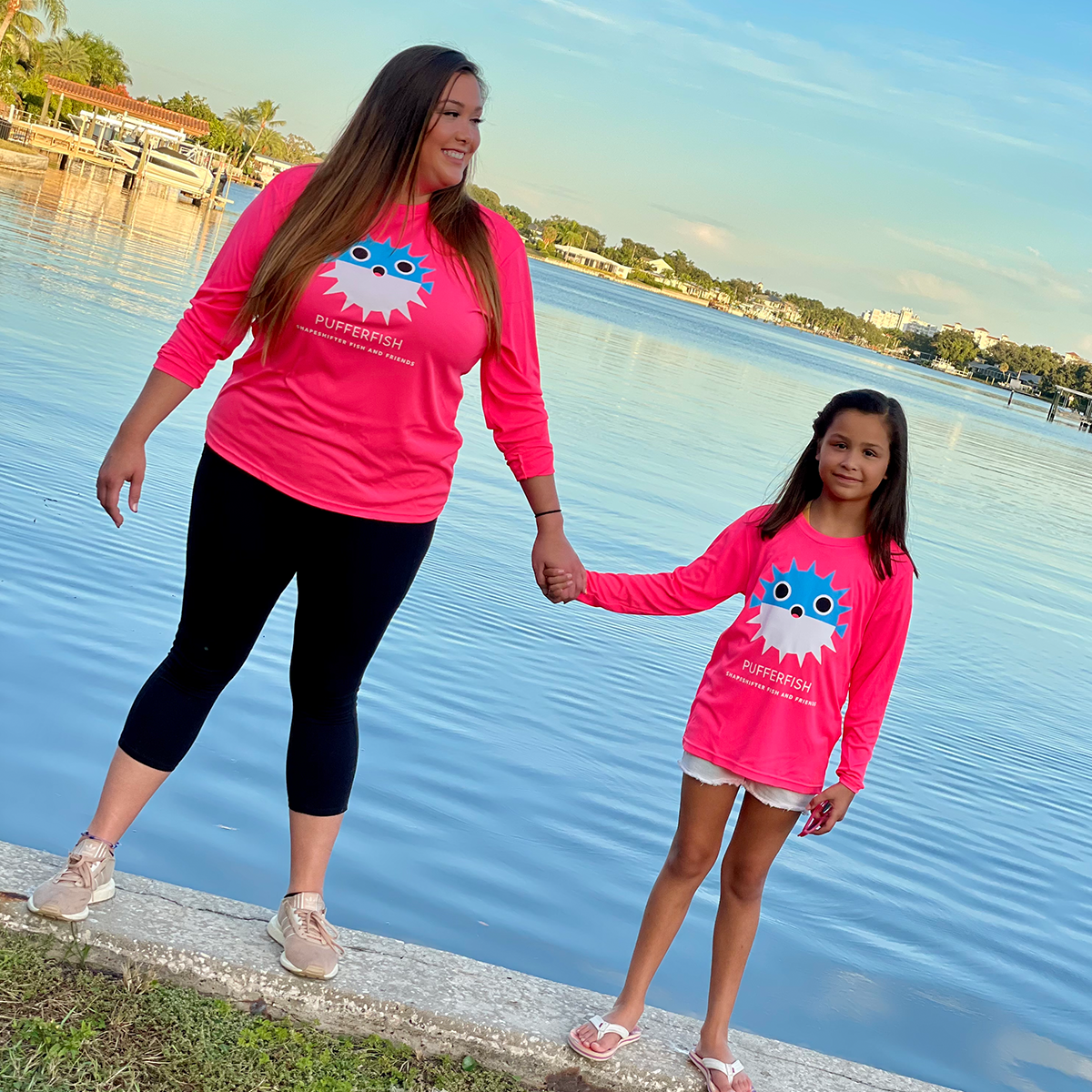 Woman and young girl holding hands by a lake, both wearing pink shirts with a blue and white logo.