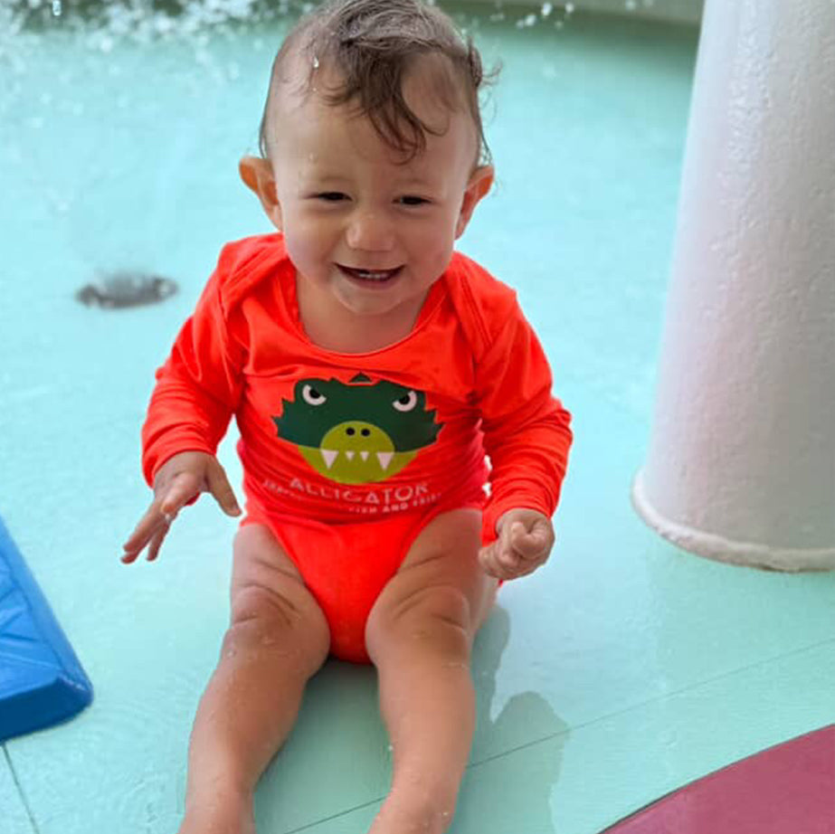 Child in an orange  Sun Protective Baby Onesie  by ShapeShifter Fish and Friends swimsuit sitting in a pool.