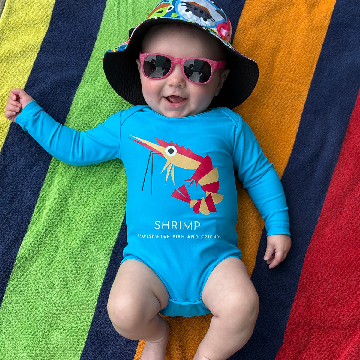 Baby wearing a Neon Blue Shrimp Upf50 Sun Protective Baby Onesie by ShapeShifter Fish and Friends, pink unbreakable sunglasses, and a colorful hat on a striped towel.