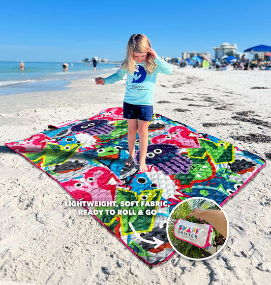 Child standing on a colorful Fish and Friends beach mat with ocean-themed design, on a sandy beach, and zoomed in shot of the beach mat rolled up