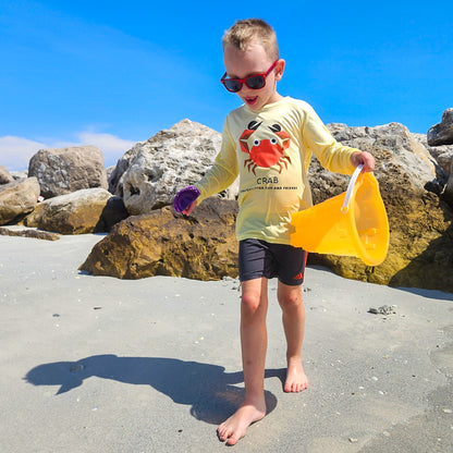 Boy playing on a beach with a yellow bucket and purple shovel, wearing unbreakable sunglasses and a Canary Yellow Crab UPF 50+ sun shirt by ShapeShifter Fish and Friends