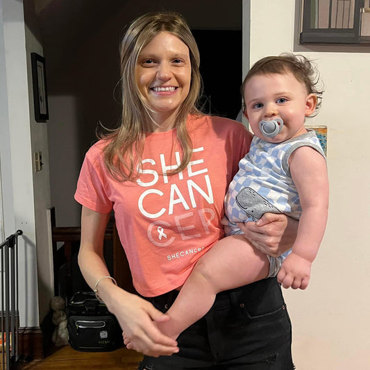 Woman holding a baby indoors wearing a 'SHE CAN' UPF cropped t-shirt by ShapeShifter Fish and Friends for breast cancer awareness lifestyle