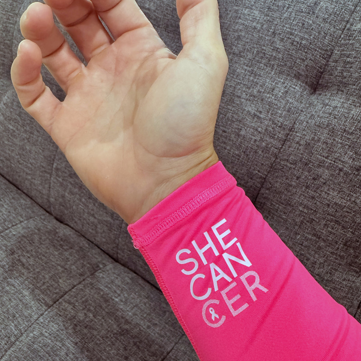 Hand wearing a pink compression sleeve with 'SHECANCER' text on a gray fabric background for breast cancer